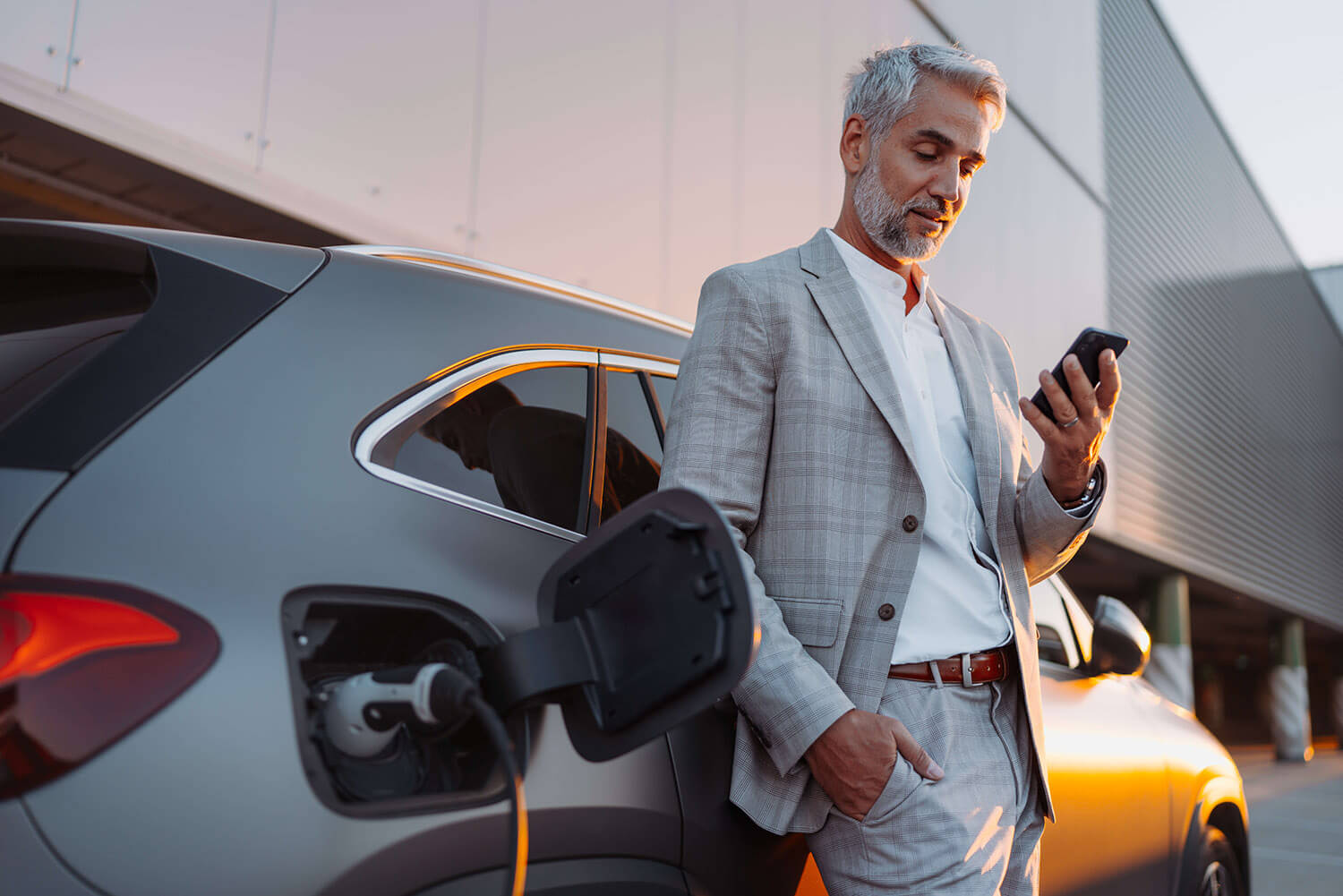 A person in a light gray suit charges a silver electric car while using a smartphone. The background shows an orange sky and modern architecture.