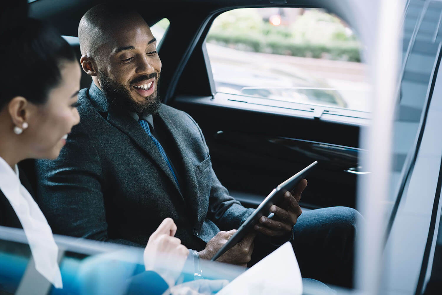 A professional dressed in a suit sitting inside a car and looking at a digital tablet, with another individual present, indicating the use of mobile technology for work purposes during transit.