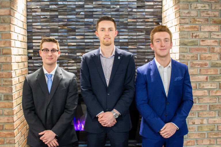 Three individuals in formal attire standing in front of a brick wall with a decorative tile feature.