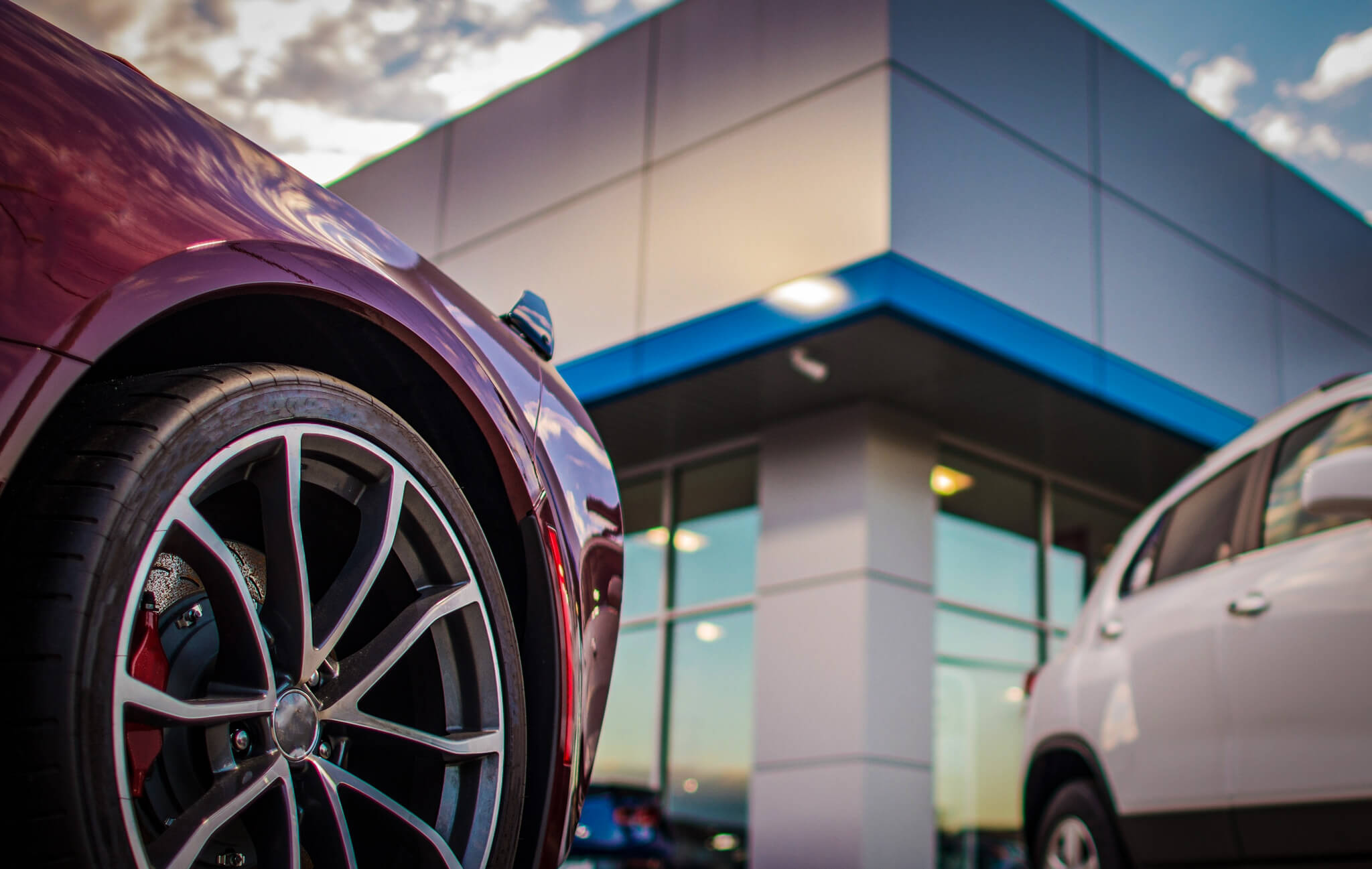 A close-up view of a car’s red front fender and silver alloy wheel with a multi-spoke design, parked outside a dealership with blue and white facade during twilight.