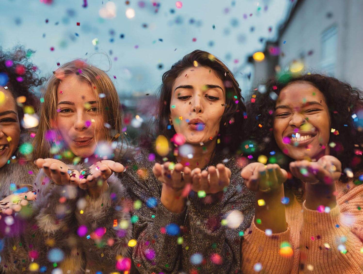 Young women blowing confetti from hands. Friends celebrating outdoors in evening at a terrace.