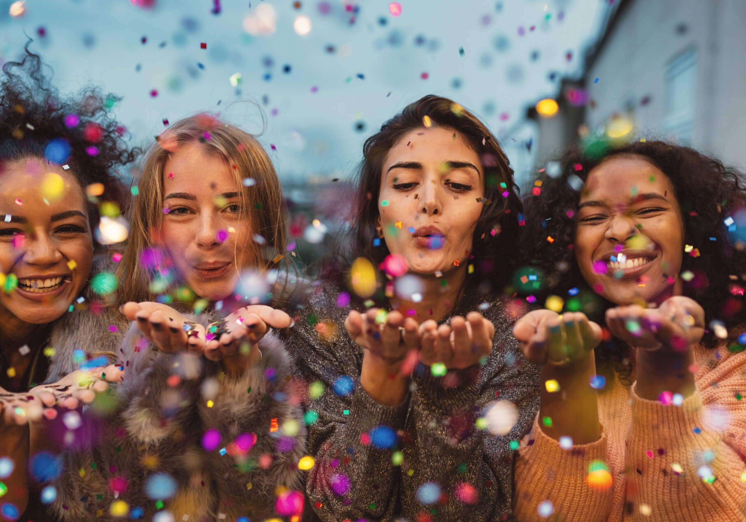 Young women blowing confetti from hands. Friends celebrating outdoors in evening at a terrace.